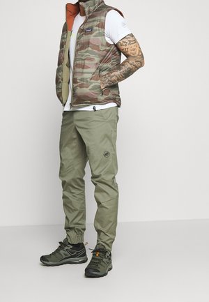 Hombre con chaleco acolchado de camuflaje, camisa blanca de manga corta, pantalones cargo verde oliva y zapatillas de senderismo verde oscuro, de pie con las manos en los bolsillos.