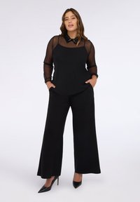 Completo nero che presenta una blusa trasparente a maniche lunghe con colletto di paillettes e pantaloni neri a gamba larga. Le scarpe sono décolleté nere a punta.