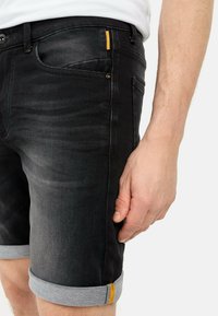 Schwarze Denim-Shorts mit umgeschlagenem Saum, hellem blauen Kontraststich und einem orangefarbenen Akzent in der Nähe der Tasche. Glatte Textur und taillierter Schnitt.