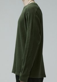 Long-sleeve, textured, dark green shirt with a ribbed pattern and round neckline, featuring a relaxed fit and straight hem.