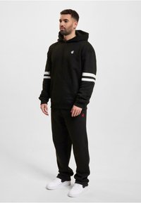 Svart hettegenser med hvite striper på ermene, kengurulomme, kombinert med svarte joggebukser og hvite sneakers. Rød logo-detalj.
