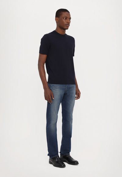 Marineblauwe t-shirt met korte mouwen, gecombineerd met lichtblauwe spijkerbroeken en zwarte schoenen. Het t-shirt heeft een ronde hals; de spijkerbroeken zijn taperend en hebben een klassieke pasvorm.