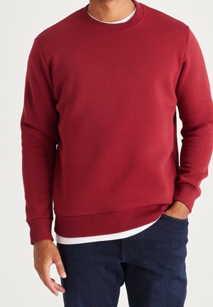 Homme portant un sweat-shirt rouge à col rond par-dessus une chemise blanche, avec un jean bleu foncé, debout devant un fond uni.