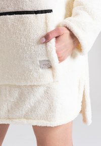 Kremowy polarowy szlafrok o puszystej fakturze, z przednią kieszenią i czarnym akcentem w postaci wstążki. Widoczna metka z logo na kieszeni.