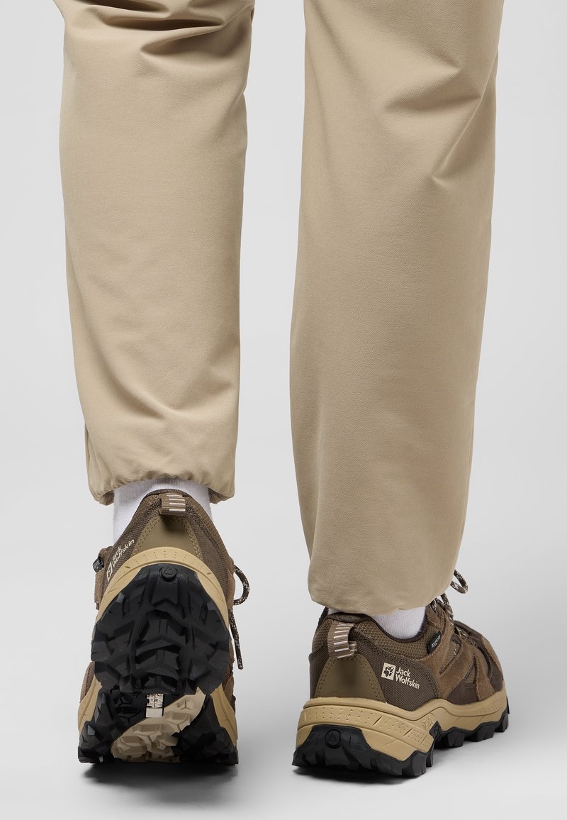 Personne portant un pantalon beige et des chaussures de randonnée marron Jack Wolfskin avec des semelles robustes, vue de dos sur un fond gris uni.