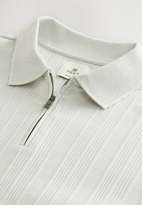 White polo shirt with a zippered collar and ribbed texture. Features a breathable mesh collar, labeled "NEXT," and slim fit design.