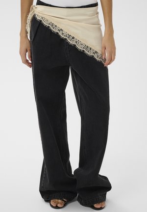 Persona che indossa jeans neri larghi, sciarpa beige con rifinitura in pizzo legata intorno alla vita, e sandali neri con cinturini su sfondo bianco.