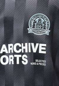 Schwarz-grau gemusteter Stoff mit weißem Text "ARCHIVE SPORTS" und einem blauen runden Aufnäher mit der Aufschrift "Perpetual Celebration of Art in Sport CORESport Archive Office."