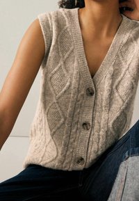 Beżowa bezrękawnik robiony na drutach z dekoltem w kształcie litery V, wzorem w warkocze i czterema guzikami z przodu. Noszona na ciemnych dżinsach.
