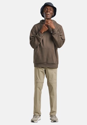 Mann trägt einen dunkelbraunen Hoodie, beige Hosen, einen Anglerhut und Turnschuhe, lächelt mit den Händen nahe dem Kinn vor weißem Hintergrund.