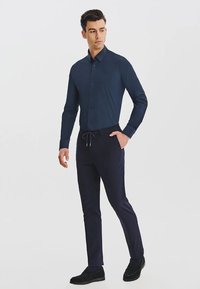 Granatowa koszula z guzikami w połączeniu z ciemnymi spodniami, z elastycznym paskiem. Model nosi czarne buty, stojąc na neutralnym tle.