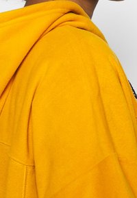 Gul huva sweatshirt i mjukt material, med en avslappnad passform och synliga sömmar. Huvan och axelkonturen synliga.