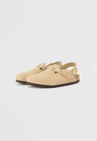 Beige mokkasiner i ruskind med justerbare spænder, rund tå og en struktureret brun sål. Flad, åben bagside for nem påtagning.