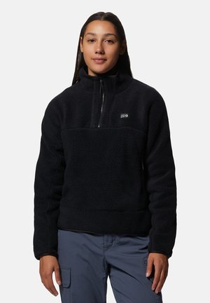 Jeune femme aux longs cheveux foncés porte un pull en polaire noir et un pantalon cargo gris, debout devant un fond clair uni.