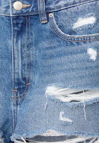 Jean en denim bleu avec des déchirures usées laissant apparaître des fils blancs, avec un bouton en métal et des coutures orange autour des poches et des coutures.