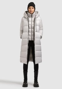 Ezüst színű steppelt kabát kapucnival, hosszú fazonú, steppelt mintázattal és elülső cipzárral. Fekete leggings és masszív fekete bakancs fölött viselve.