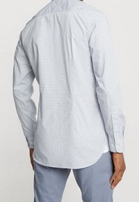 Mężczyzna w jasnoniebieskiej koszuli w kratę z długim rękawem i jasnoniebieskich spodniach, widoczny od tyłu, stojący na tle jednolitym.