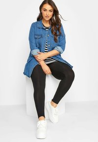 Jeansowa kurtka z podwiniętymi rękawami, paski w biało-czarnym kolorze na bluzce, czarne legginsy i białe trampki, siedząca na białym pedestal.