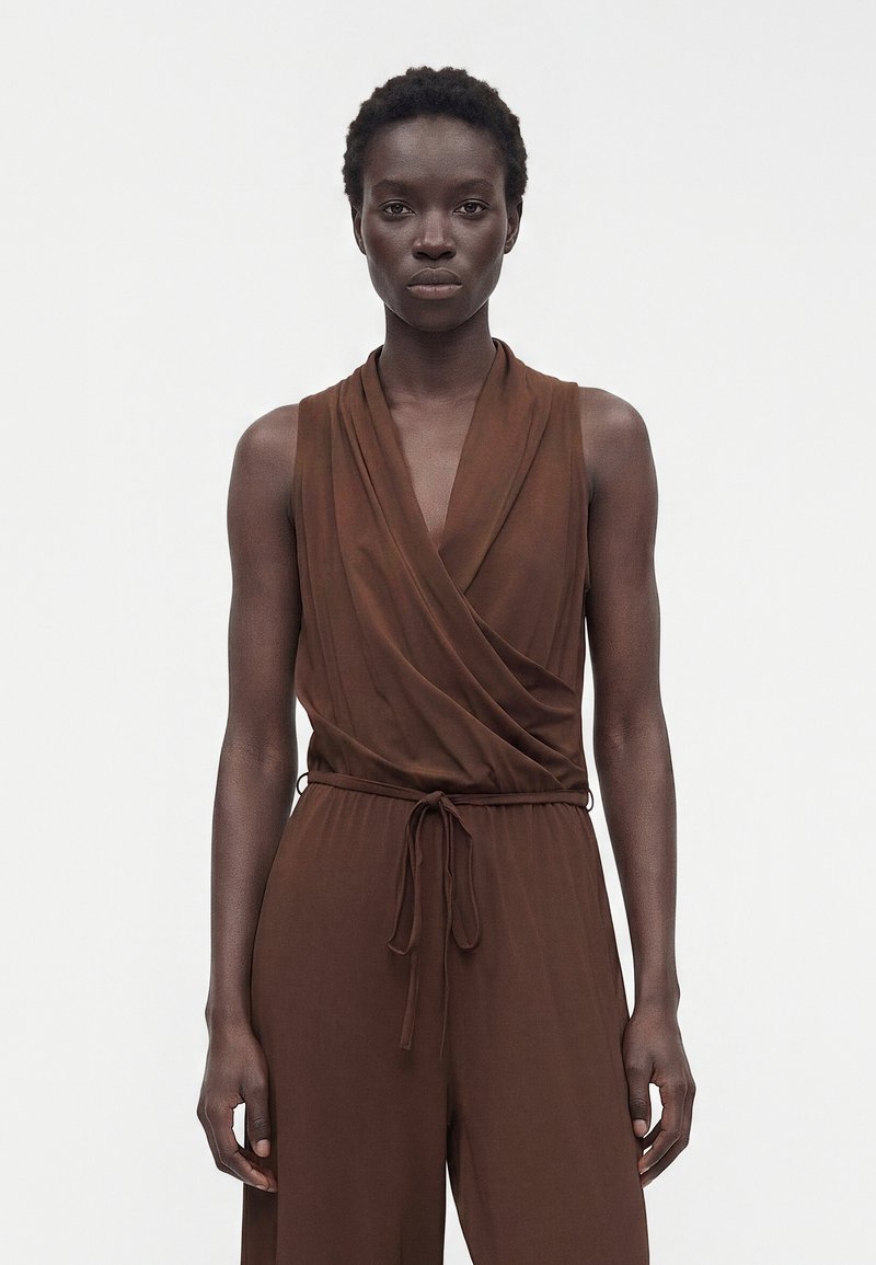 Femme aux cheveux courts et bouclés portant une combinaison marron sans manches style cache-cœur avec une taille à cordon, debout devant un fond uni.