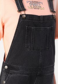 Zwarte denim tuinbroek met verstelbare schouderbanden, borstzak en metalen accenten, gecombineerd met een lichtroze t-shirt met grafisch ontwerp.