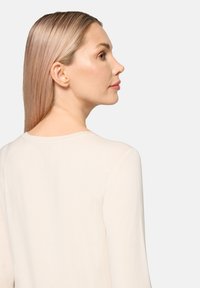 Długi jasnobeżowy top z długim rękawem, okrągłym dekoltem, gładką fakturą i minimalistycznym wzorem. Modelka pokazana od tyłu, włosy proste i jasne.