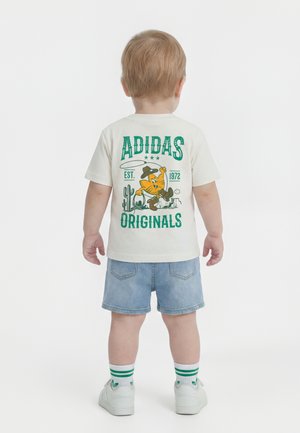 Παιδί προσχολικής ηλικίας, γυρισμένο πλάτη, φοράει λευκό t-shirt Adidas με καρτούν θεματολογία καουμπόη, τζιν σορτς, λευκά αθλητικά παπούτσια και ριγέ κάλτσες.