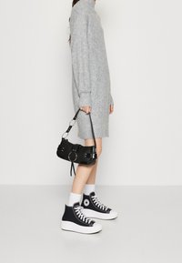 Grå stickad klänning med långa ärmar, svart lackhandväska med cirkulärt hårdvara, vita och svarta högstlupna sneakers, och vita ankelsockor.