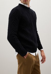 Uomo con un maglione blu navy a trecce sopra una camicia bianca e pantaloni marroni, con le mani nelle tasche, su uno sfondo chiaro e uniforme.