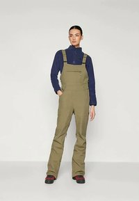 Oliivroheline kombinesoon reguleeritavate õlapaeltega, lukuga taskud ja kitsenev jala disain, koos tumesinise tõmblukuga pluusiga.