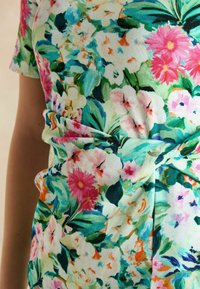 Blumenkleid mit einem hellgrünen Hintergrund, lebendigen pinken und blauen Blumen, kurzen Ärmeln und einem geknoteten Design auf Taillenhöhe.