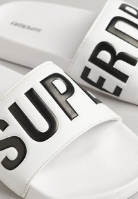 Slidesandaler med en hvid tekstureret bund og en bred sort rem med ordet "SUPER" i fed, hævet skrift.