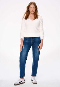 Witte, fluffy V-hals trui gecombineerd met blauwe spijkerbroek. Witte sneakers met zwarte strepen. Model staat tegen een effen achtergrond.