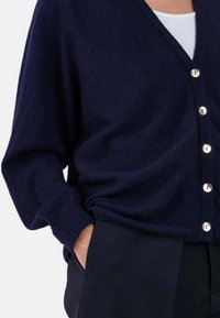 Cardigan bleu marine, texture douce, col en V, grands boutons, coupe décontractée avec épaules tombantes, associé à un pantalon foncé. Convient pour le superposition.