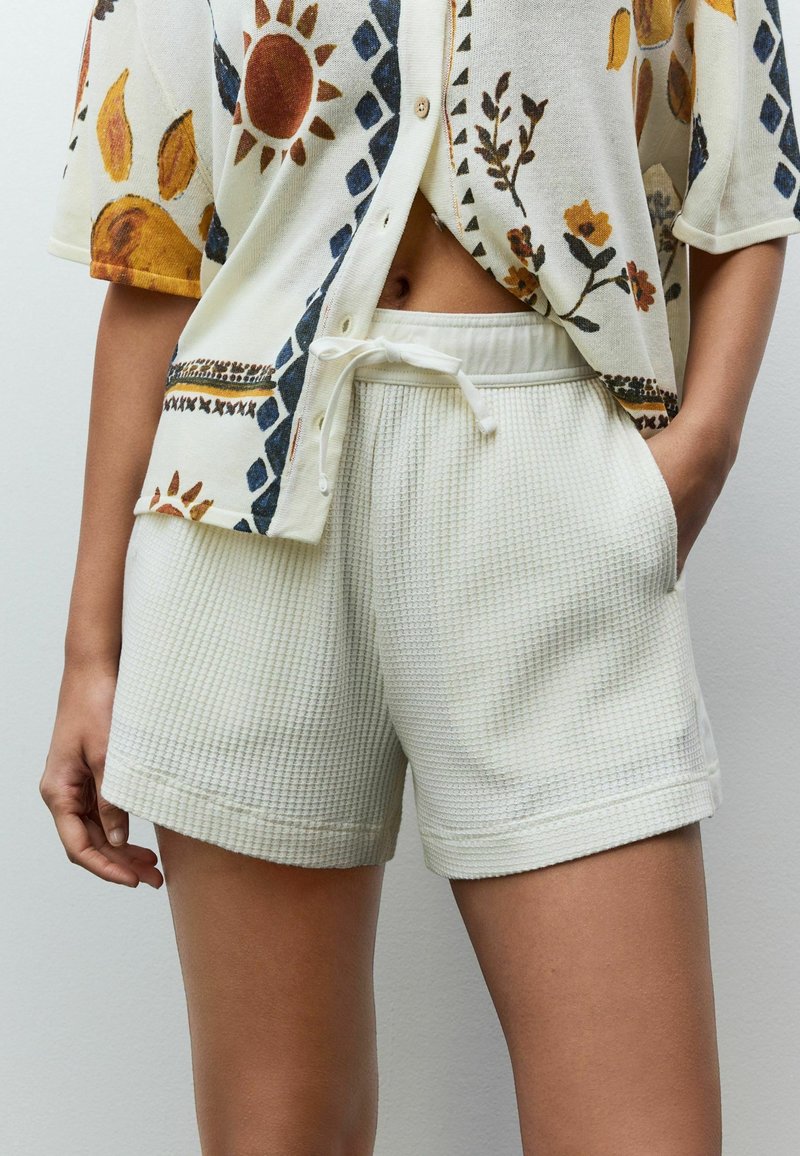 Femme portant un short blanc texturé avec cordon de serrage et une chemise boutonnée avec des motifs soleil et fleurs, main reposant dans la poche du short.