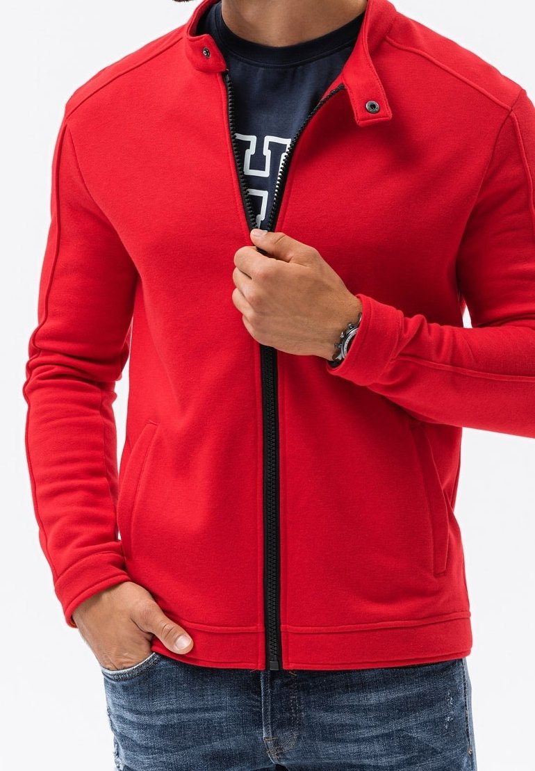 Giacca rossa con zip in tessuto morbido, caratterizzata da un colletto alto, tasche laterali e dettagli in zip nera, indossata sopra una t-shirt blu navy.