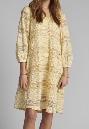 Femme portant une robe ample jaune clair à manches trois-quarts avec de délicats motifs en dentelle ajourée, debout devant un fond uni.