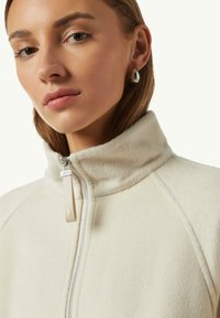 Giacca zip-up color avorio chiaro realizzata in tessuto morbido e testurizzato con colletto alto e zip argento. Presenta un'etichetta rettangolare in tessuto per la chiusura.