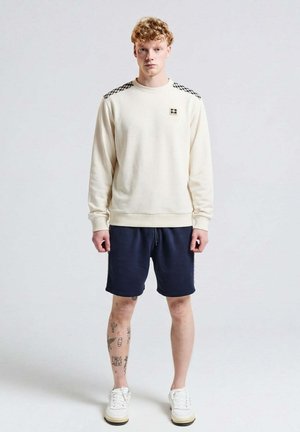 Giovane con capelli ricci che indossa una felpa crema con spalle a quadri, pantaloncini blu navy, scarpe da ginnastica bianche e tatuaggi sulle gambe, in piedi contro uno sfondo semplice.