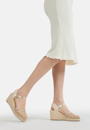 Gambe con gonna midi bianca e sandali beige intrecciati con zeppa e cinturini alla caviglia su sfondo bianco.