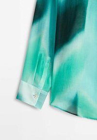 Manche et poignet d'une chemise à manches longues avec un dégradé de couleurs bleu sarcelle et aqua, présentant un seul bouton beige sur le poignet.