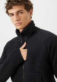 Giovane uomo che indossa una giacca in pile nera con superficie strutturata e maniche con foro per il pollice, tiene la cerniera vicino al petto su uno sfondo neutro.