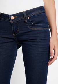Jeans in denim scuro con un design aderente, cuciture arancioni a contrasto, tasche anteriori e un bottone in rame in vita.