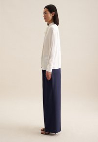 Hvid langærmet bluse med høj krave, parret med marineblå brede bukser. Glat stofstruktur, minimalistisk design og flade sandaler.