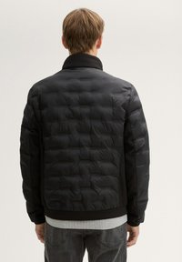 Černá péřová bunda s prošitým vzorem, žebrovaným límcem a černými rukávy, doplněná šedou vrstvenou košilí pod ní.