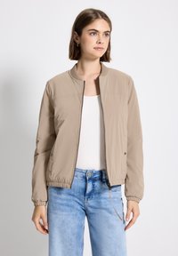 Jeune femme aux cheveux bruns courts portant une veste bomber beige à fermeture éclair, un t-shirt blanc et un jean bleu clair, se tenant devant un fond blanc.