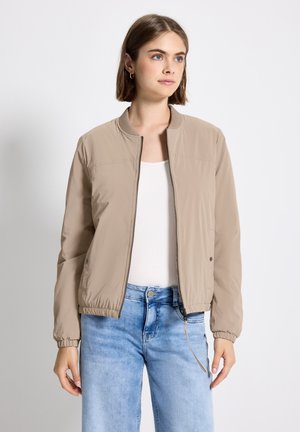 Jeune femme aux cheveux bruns courts portant une veste bomber beige à fermeture éclair, un t-shirt blanc et un jean bleu clair, se tenant devant un fond blanc.