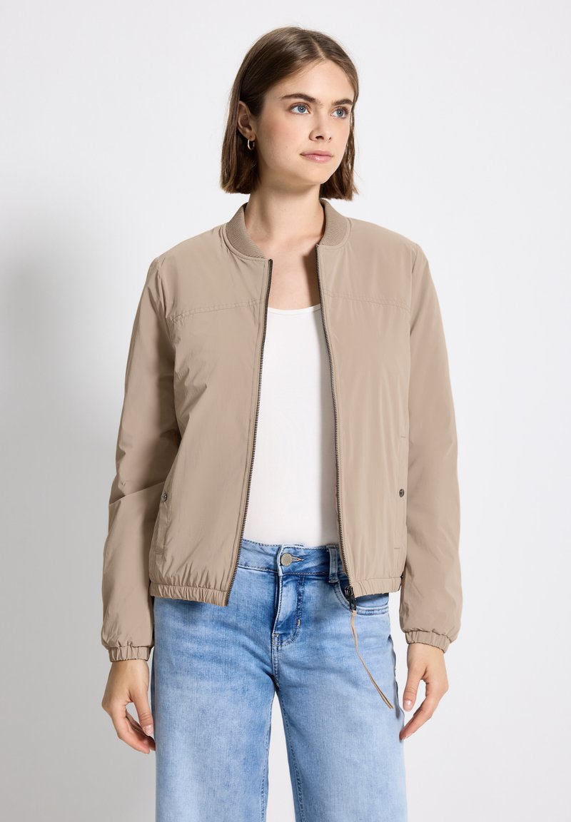 Jeune femme aux cheveux bruns courts portant une veste bomber beige à fermeture éclair, un t-shirt blanc et un jean bleu clair, se tenant devant un fond blanc.