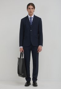 Abito blu navy con vestibilità slim, giacca a un pulsante, abbinato a una camicia bianca e una cravatta a righe. Tiene una borsa a tracolla in pelle nera, indossando scarpe lucide nere.