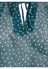 Türkise Bluse mit weißen Punkten, mit einem Ausschnitt in Schlaufenform und Bindebändern. Leichter, strukturierter Stoff verstärkt das Design.