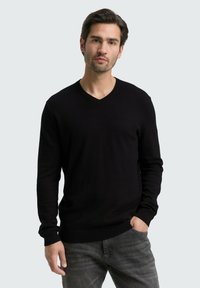 Schwarzer V-Ausschnitt-Pullover in gerippter Textur, mit langen Ärmeln und taillierter Passform, kombiniert mit dunkelgrauen Jeans. Schlicher Hintergrund.